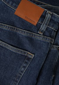 Vaqueros de mezclilla oscura con un parche de cuero marrón etiquetado "TNTUUM", detalles de costura visibles y un diseño clásico de cinco bolsillos.