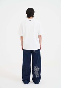 T-shirt bianca a maniche corte con collo rotondo, abbinata a jeans in denim blu larghi con un design di drago sulla gamba sinistra.