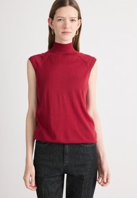 Top rosso senza maniche in un morbido tessuto lavorato a maglia, con una forma aderente e colletto a costine. Abbinato a jeans grigi scuri a gamba dritta.
