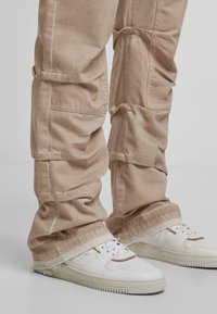 Beige cargo-housut, joissa on kootut polvet, valkoisten nauhallisten sneakerien päällä, joissa on vaaleanruskeita yksityiskohtia, seisoen yksinkertaisella taustalla.