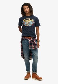 Camiseta de algodón azul marino con gráfico vintage, diseño en azul y amarillo. Jeans azul claro, camisa de cuadros roja y negra atada a la cintura, botas marrones.