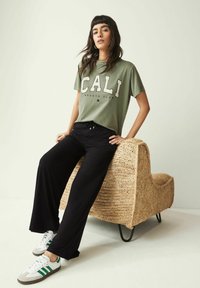 Militærgrøn grafisk t-shirt med "CALI" tekst, parret med sorte brede bukser og hvide sneakers med grønne striber, siddende på en flettet stol.