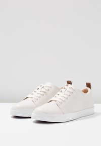 Valkoiset nahkakengät, joissa on pyöreät varpaat, nauhakiinnitys edessä, kontrastivärinen vaalea kantapääkappale ja valkoiset kumipohjat. Liukas pinta, minimalistinen muotoilu.