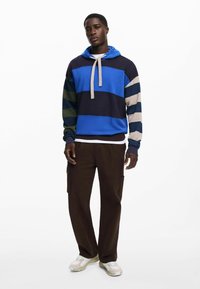 Hooded sweatshirt featuring horizontal stripes in blue, navy, cream, and green, with a drawstring and ribbed hem. Paired with brown cargo pants.