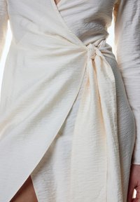 Helles beiges Wickelkleid mit strukturierter Oberfläche, das ein Knoten-Detail an der Taille und lange Ärmel aufweist, was einen asymmetrischen Saum erzeugt.