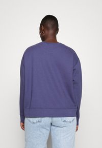 Persona con capelli corti che indossa orecchini a cerchio argento, una felpa larga di un viola scuro e jeans azzurri, vista di spalle su uno sfondo bianco.