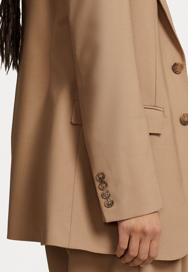 Beige taillierter Blazer mit Reverskragen, vier braunen Knöpfen am Ärmel und einem glatten, strukturierten Stoff.