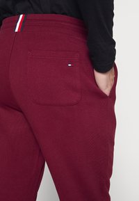 Burgunderfarbene Sweatpants haben einen flachen, elastischen Bund mit einem farbigen Streifenakzent und einer einzigen Gesäßtasche. Glatte Stoffstruktur.