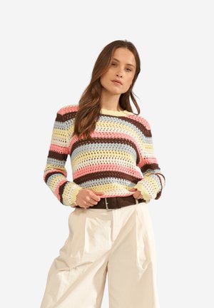 Femme portant un pull crocheté à rayures pastel rose, marron, jaune et bleu, rentré dans un pantalon crème taille haute avec une ceinture sombre.