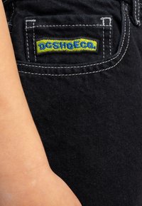 Jeans de mezclilla negra con un bolsillo que presenta el logo bordado "DC SHOES" en amarillo y azul. Costuras en contraste a lo largo de los bordes. Textura suave.