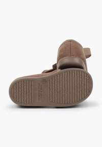 Brune ruskindsko med rund tå, elastisk rem og tekstureret gummisål med en bølget struktur. Sålen viser etiketten "Made in Spain".