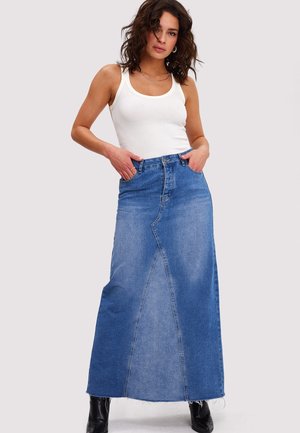 Vrouw draagt een wit mouwloos topje, een lange blauwe denim rok met een naad aan de voorkant en zwarte enkellaarzen, staand met handen in de zakken.