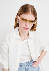 Lunettes de soleil teintées en jaune avec un design élégant et des accents décoratifs en forme d'aile. Portées avec une chemise blanche à boutons et un jean bleu clair.