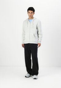 Grå hettegenser med glidelås, hette, frontlommer og logodetalj. Kombinert med svarte bukser og grå sneakers. Glatt tekstur, uformell passform.