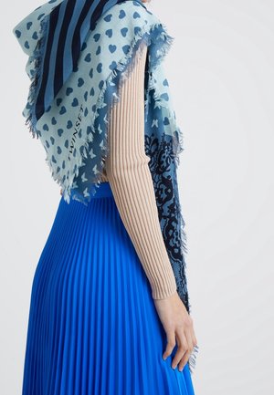 Écharpe bleue à motifs de cœurs et de rayures, bords frangés, drapée sur un haut beige à manches longues côtelé, associée à une jupe plissée bleue.