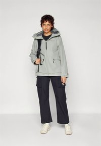 Lysegrå vandtæt jakke med hætte, lynlåsføring foran og to sidelommer. Båret med sorte cargobukser og hvide sneakers.