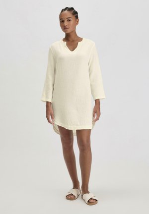 Weißes, strukturiertes Kleid mit V-Ausschnitt und Dreiviertelärmeln, das einen lockeren Schnitt und einen leicht längeren Saum hinten aufweist.