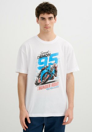 Joven con camiseta blanca con gráfico azul de motocicleta y texto "Speed Demon 95 Thunder Road" de pie contra un fondo liso.