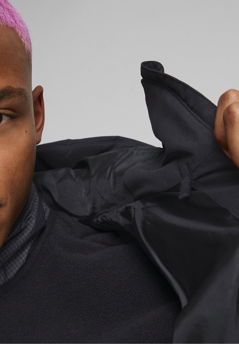 Vêtement d'extérieur noir avec une texture douce, doté d'un col zippé et d'une doublure lisse. Main visible tenant le col, mettant en valeur les couches de tissu.