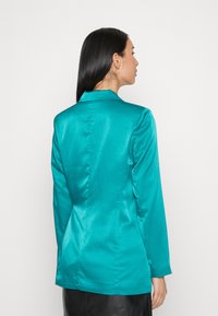 Blazer en satin teal avec un design ajusté, à fermeture simple sur le devant et manches longues. Doté d'un col cranté et d'une texture lisse.