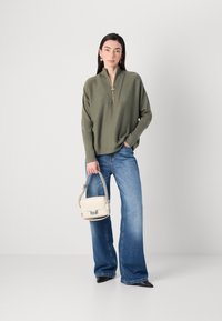 Eine Frau steht selbstbewusst da, trägt einen grünen Reißverschluss-Pullover und eine blaue Jeans und hält eine kleine cremefarbene Handtasche. Ihre elegante Styling ergänzt ihr Erscheinungsbild.
