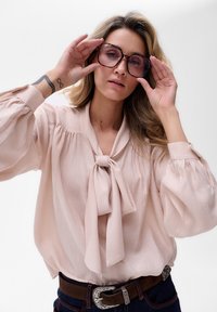 Jasnoróżowa bluzka z kokardą przy kołnierzyku, długimi bufkami i fakturowanym materiałem; zestawiona z ciemnym dżinsem i paskiem z srebrną klamrą.