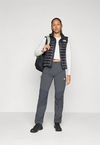 Mustast isoleeritud vest valge pika varrukaga särgi peal, mille juurde on sobitatud hallid püksid tumedamate paneelidega. Modell hoiab musta seljakotti.