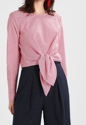 Kobieta w czerwono-białej, pionowo paskowanej bluzce z długim rękawem, związaną w talii, w połączeniu z ciemnymi spodniami w pinstripe o wysokim stanie.