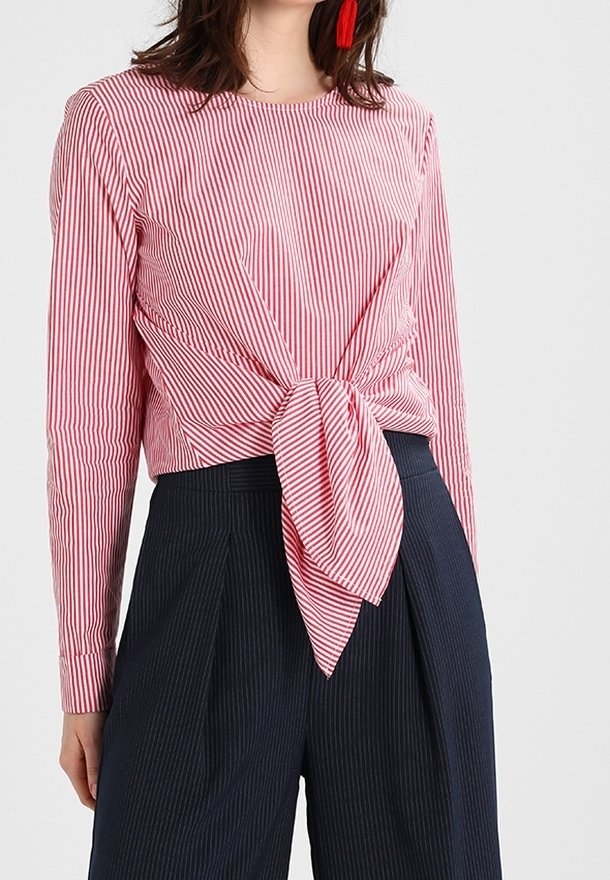 Femme portant un haut à manches longues rayé verticalement rouge et blanc noué à la taille, associé à un pantalon taille haute à fines rayures foncé.