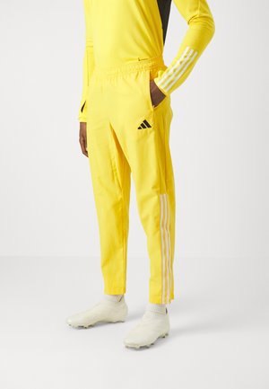 Person trägt leuchtend gelbe Adidas-Trainingshose mit weißen Streifen, Hand in der Tasche, weiße Fußballschuhe und passendes gelbes Langarmoberteil.