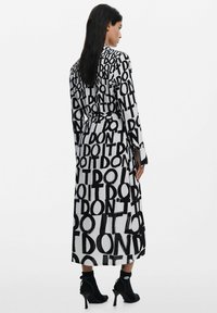 Schwarzes und weißes langes Kleid mit einem auffälligen, grafischen Textmuster "WIEDER NICHT TUN". Ausgestellte Ärmel und ein verstellbarer Gürtel werten das Design auf.
