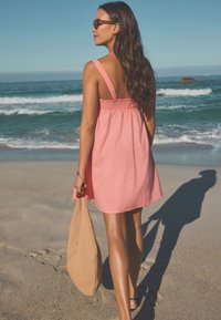 Ljusrosa ärmlös klänning med rynkad rygg och breda axelband. Bärs med en brun texturerad väska, på en strand med vågor i bakgrunden.