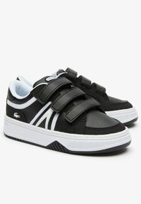 Sneakers Lacoste noir et blanc avec trois bandes velcro, cuir texturé et semelles en caoutchouc blanc, inclinés sur un fond blanc.