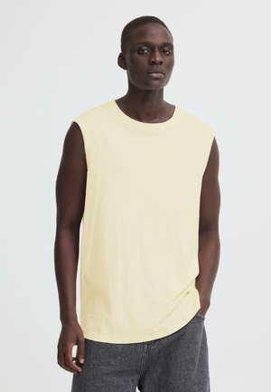 Young Black man wearing a sleeveless pale yellow shirt and dark gray pants standing against a white background.