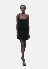 Schwarzes ärmelloses Kleid mit einem geschichteten Oberteil und ausgestelltem Rock, aus strukturiertem Stoff gefertigt. Getragen mit flachen Sandalen.
