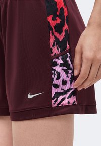 Μπορντό αθλητικό σορτς με ροζ και κόκκινα πάνελ δίχτυ παραλλαγής στα πλάγια, λογότυπο Nike κοντά στο στρίφωμα και το χέρι ενός ατόμου που ακουμπά δίπλα στον μηρό.