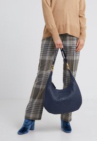 Marinblå läderhandväska med en texturerad yta och präglad logotyp, med en kurvad form och avtagbart band. Kombineras med rutiga byxor.