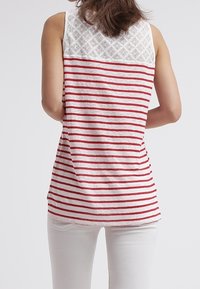 Žena v bezrukávovém bílém topu s červenými vodorovnými pruhy a krajkovým detailem na horní části zad, v kombinaci s bílými kalhotami.