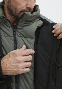 Zunanja jakna v črni barvi z zeleno podlogo. Ima visok ovratnik in dvojne zadrge. Vidna teksturirana tkanina in šivi.