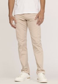 Pantalon en coton beige avec une coupe slim et une texture subtile, associé à des baskets blanches avec un accent doré. La chemise est blanche unie.