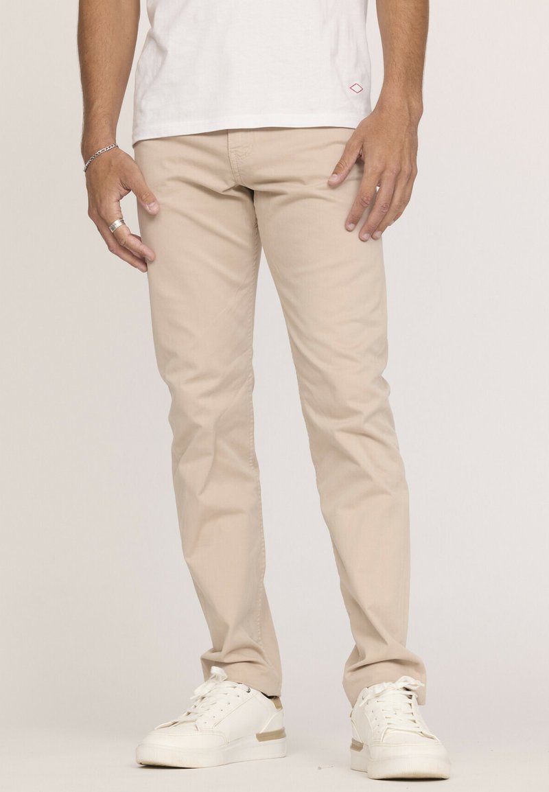 Pantalon en coton beige avec une coupe slim et une texture subtile, associé à des baskets blanches avec un accent doré. La chemise est blanche unie.