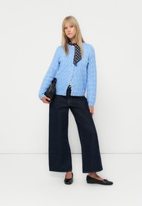 Vaaleansininen kaarrokkeellinen neuletakki pyöreillä nappeilla, yhdistetty tummiin leveälahkeisiin denim-farkkuihin ja mustiin ballerinatossuihin, asusteena pilkullinen huivi.