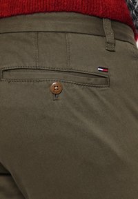 Olivgrüne Hose aus weicher Textur mit einer einzelnen Gesäßtasche, die mit einem braunen Knopf und einem kleinen dreifarbigen Logo-Element versehen ist.