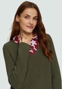 Olivgrüner gestrickter Pullover mit V-Ausschnitt, über einem kastanienbraunen Hemd mit Kragen, das ein hellrosa Blumenmuster zeigt.