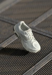 Białe buty do biegania Nike z siateczkowym i syntetycznym wykończeniem spoczywające na metalowej kratce podłogowej w miękkim naturalnym świetle.