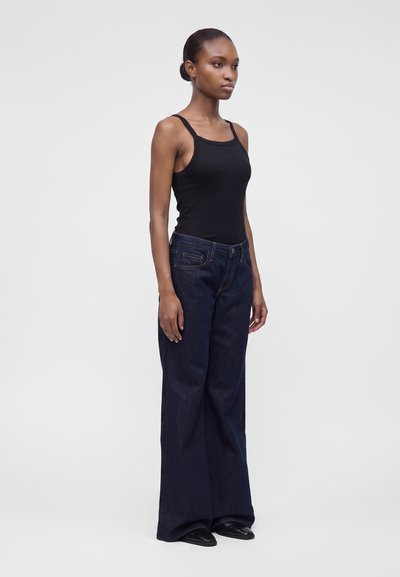 Jeune femme debout légèrement tournée vers la droite, portant un débardeur noir et un jean bleu foncé ample avec des chaussures noires sur un fond uni.