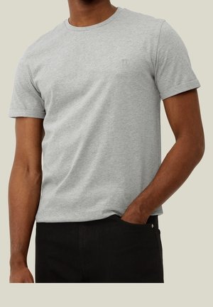 Man wearing a plain light gray short-sleeve t-shirt with a small chest logo and black pants, one hand in pocket.