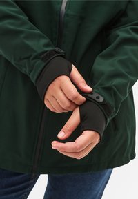 Donkergroene softshell jas met zwarte elastische manchetten. Voorzien van een ritssluiting aan de voorkant, klittenband accenten en een lichte textuur. Geschikt om in laagjes te dragen.