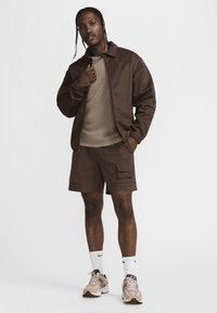 Brun letvægtsjakke over en beige skjorte, parret med brune cargo shorts. Hvide strømper og beige atletik sko fuldender outfittet.