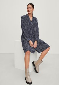 Vestito camicia a maniche lunghe di colore blu navy con un motivo di foglie, caratterizzato da un colletto a aventer e una vestibilità ampia, abbinato a stivaletti alla caviglia in paglia beige.